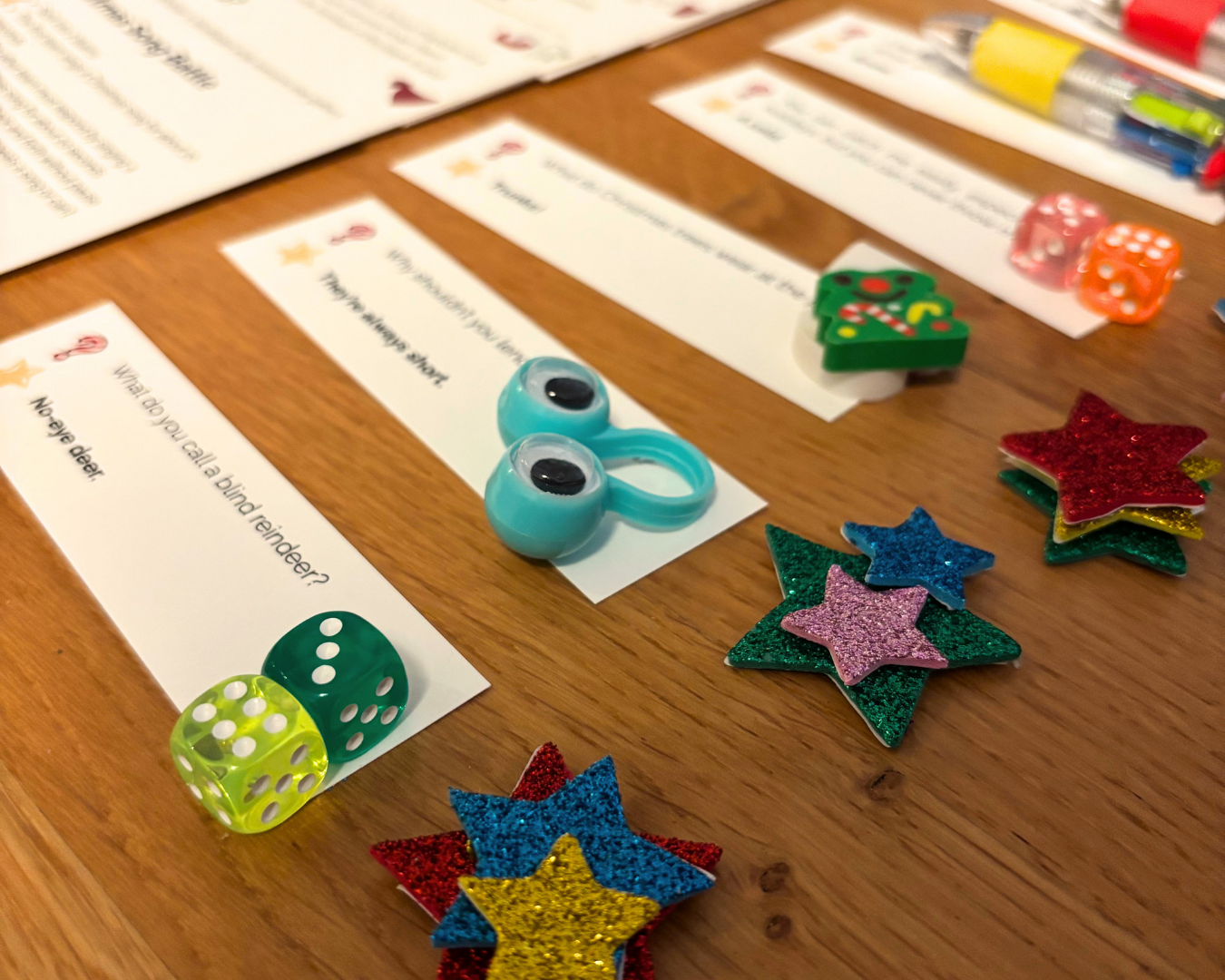 A close-up of a wooden tabletop with several small printed cards, colorful dice, a turquoise eye ring, and glittery star cutouts in red, blue, green, yellow, and pink.
