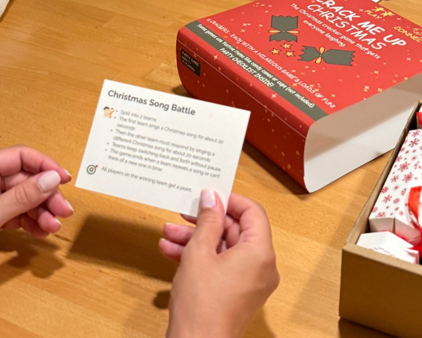 A person's hands hold a white instruction card titled 'Christmas Song Battle' in front of a red gift box labeled 'Crack Me Up Christmas' on a wooden table.