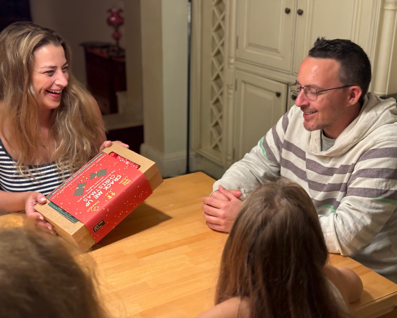 A family shares a joyful moment around a wooden table, with a woman holding a red Crack Me Up Christmas game box labeled and a man in a striped hoodie smiling nearby.