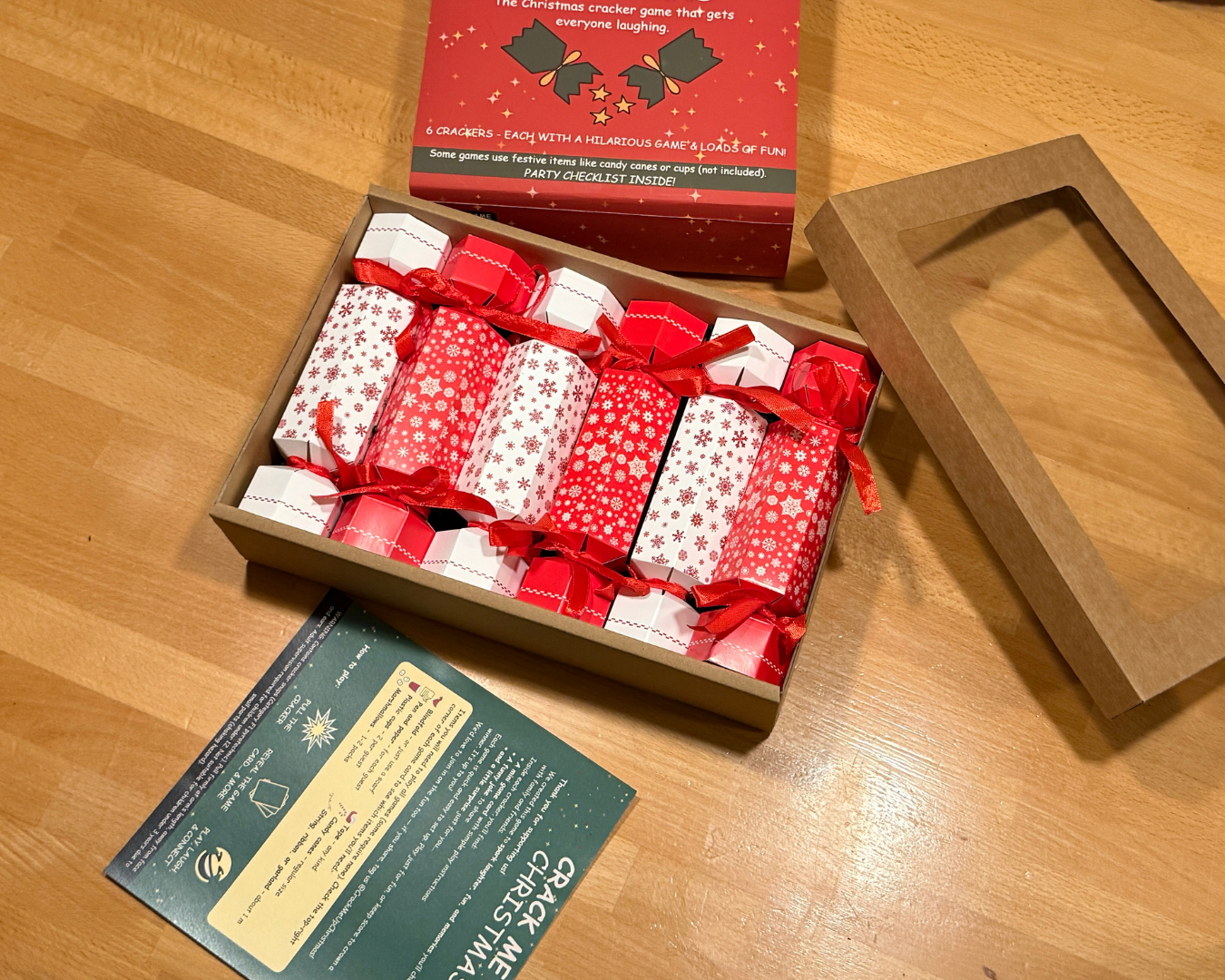 A boxed set of Christmas cracker game muffs arranged in an open kraft cardboard tray on a wooden tabletop. The muffs are wrapped in red and white patterned paper with red ribbons.